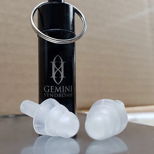 Two translucent silicone earplugs displayed in front of a black cylindrical tube labeled 'GEMINI SYNDROME' with a silver key ring, on a reflective surface against a cardboard background.