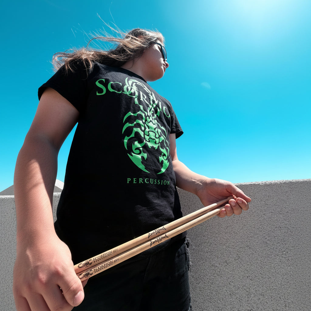 Person wearing a black t-shirt with green scorpion percussion logo and text, holding drumsticks against a clear blue sky.